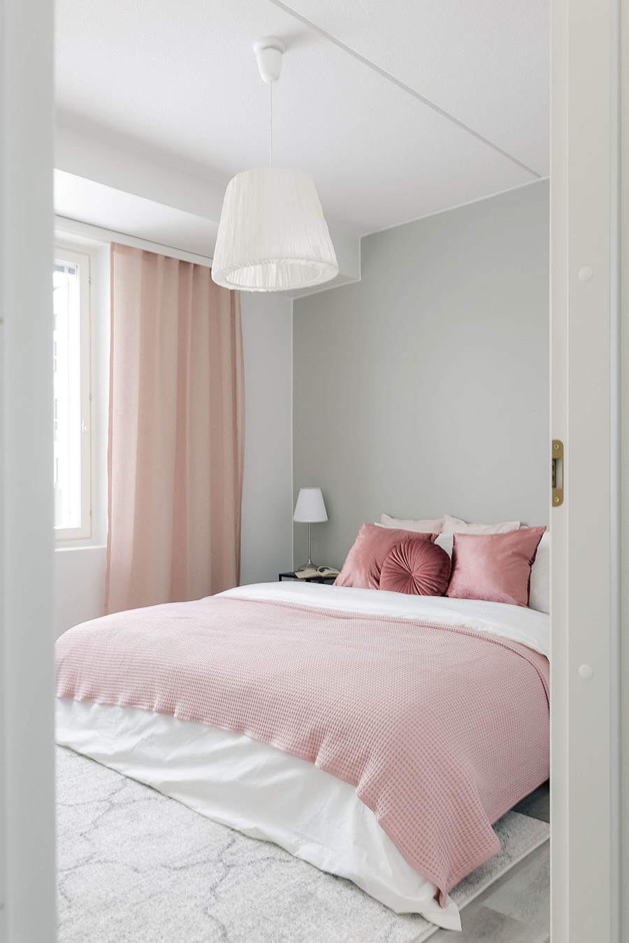 Valitsemalla värikkäitä tekstiilejä, saat vaaleaankin kotiin väriä. Kuvassa vaaleaan makuuhuoneeseen on pedattu vaaleat petivaatteet ja vaalenpunainen päiväpeitto sekä tyynyjä. Ikkunassa on vaalenpunaiset verhot.